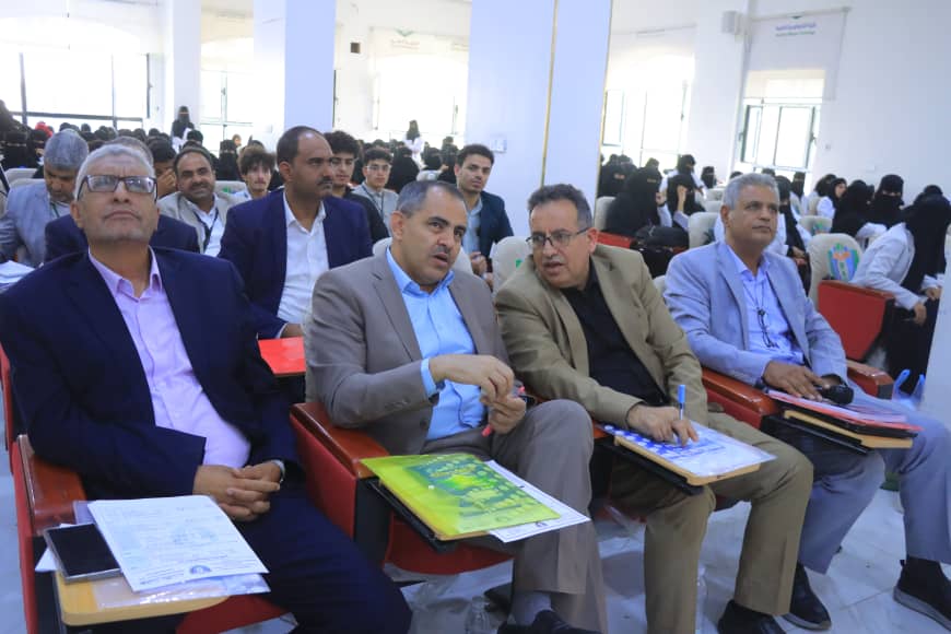 Four men in suits sit in the front row of a bright conference hall, with notebooks and folders on their desks.