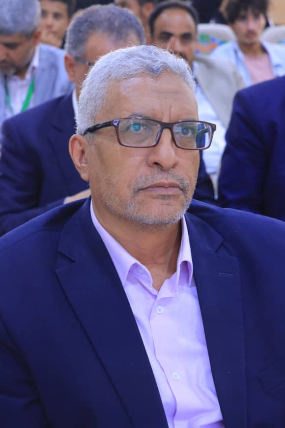 Man with gray hair and glasses in a navy suit and light purple shirt seated at a conference, with people in the background.