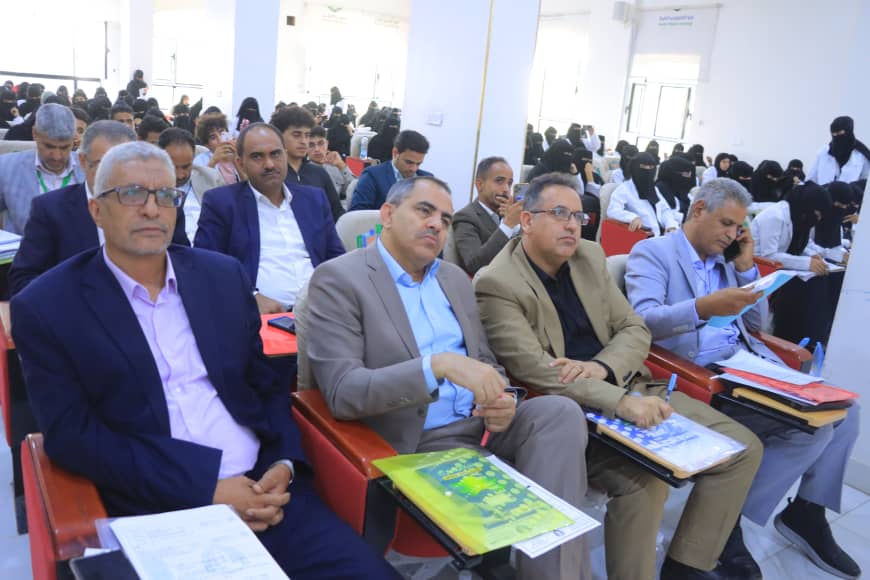 Audience seated in a bright conference hall, foreground: five men in suits with folders and papers. Behind them, rows of attendees, many women in black hijabs.