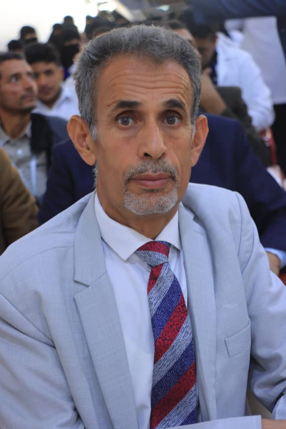 Middle-aged man in a light gray suit and striped red-blue tie at a formal event, looking directly at the camera with a neutral expression.