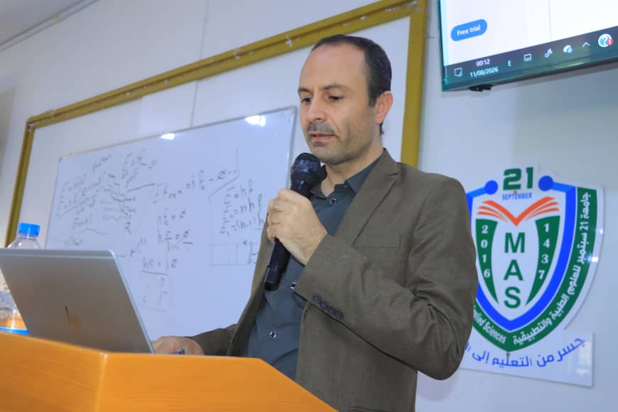 A man in a brown blazer speaks into a microphone at a podium with a laptop, a whiteboard, and a MAS banner in the background.