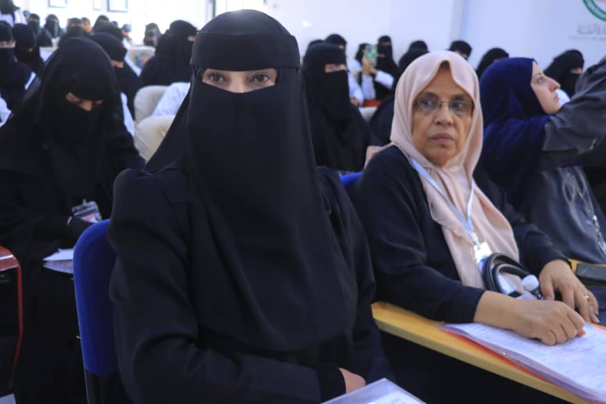 Front-and-center woman in a black niqab seated at a desk in a conference room, with other women in hijabs behind her, listening to a presentation.