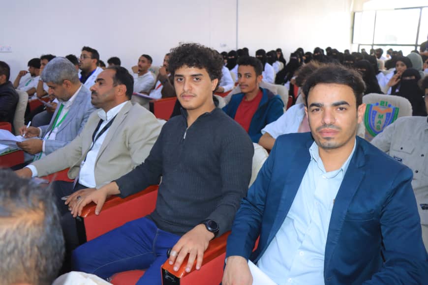 Front-row attendees at a conference, with two men facing the camera while others sit behind them in the auditorium.
