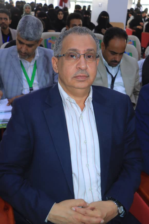 Middle-aged man wearing glasses and a navy suit sits with hands folded at a conference, with attendees visible in the background.