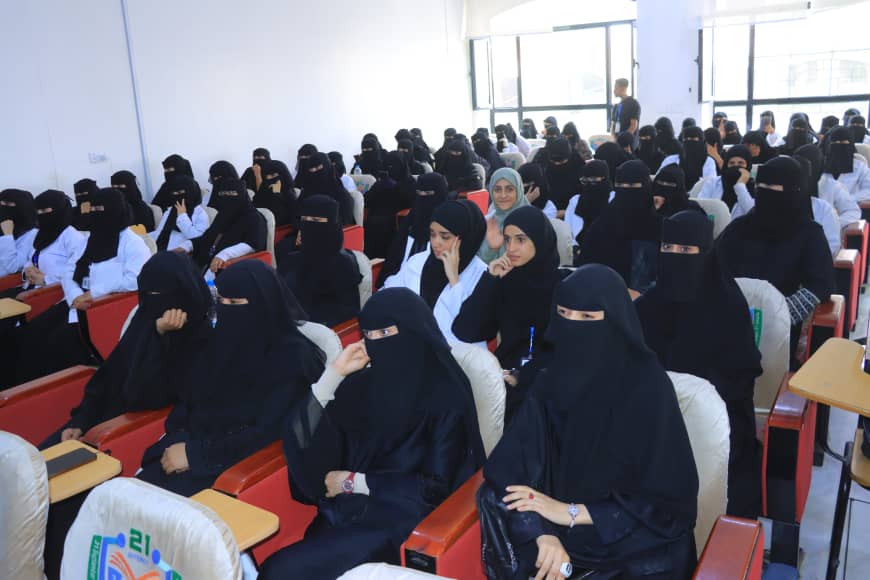 A large classroom filled with women wearing niqabs attending a lecture in a lecture hall with red chairs and windows in the back.
