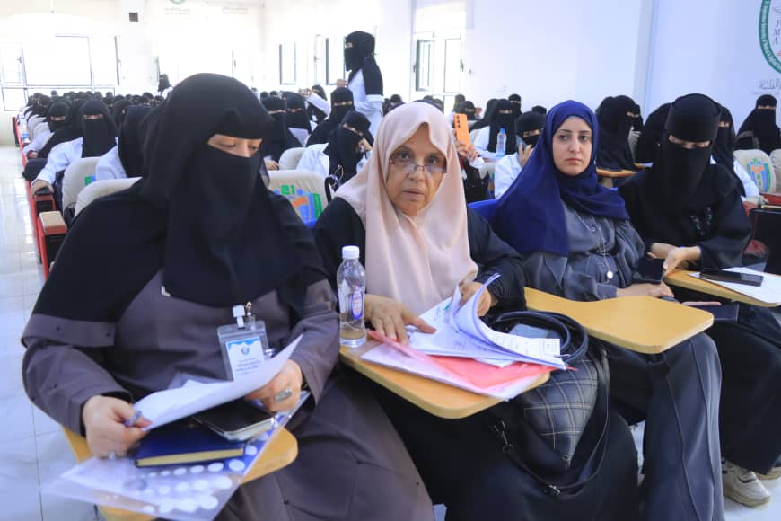 Several women wearing hijabs seated in a bright classroom, taking notes with papers and folders.