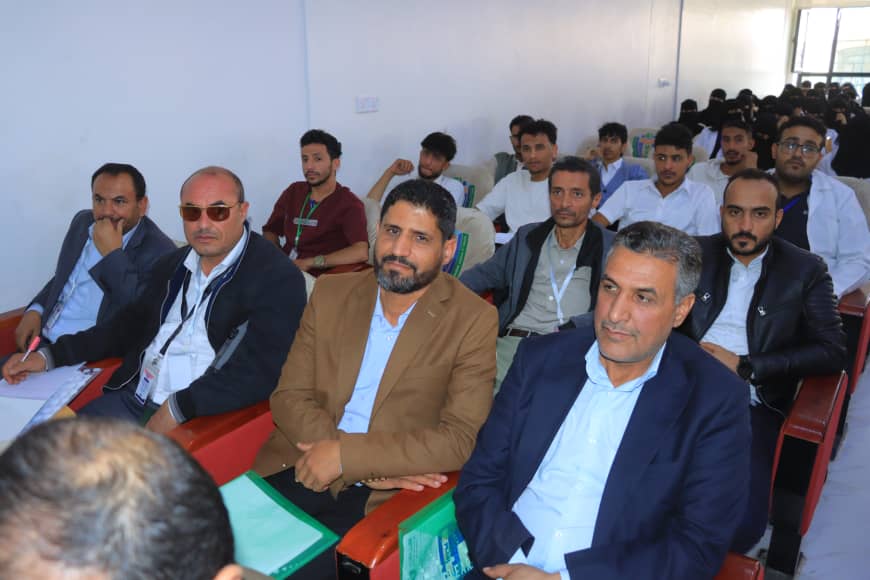 Group of men seated in a conference room, front rows in suits with lanyards, listening to a presentation.