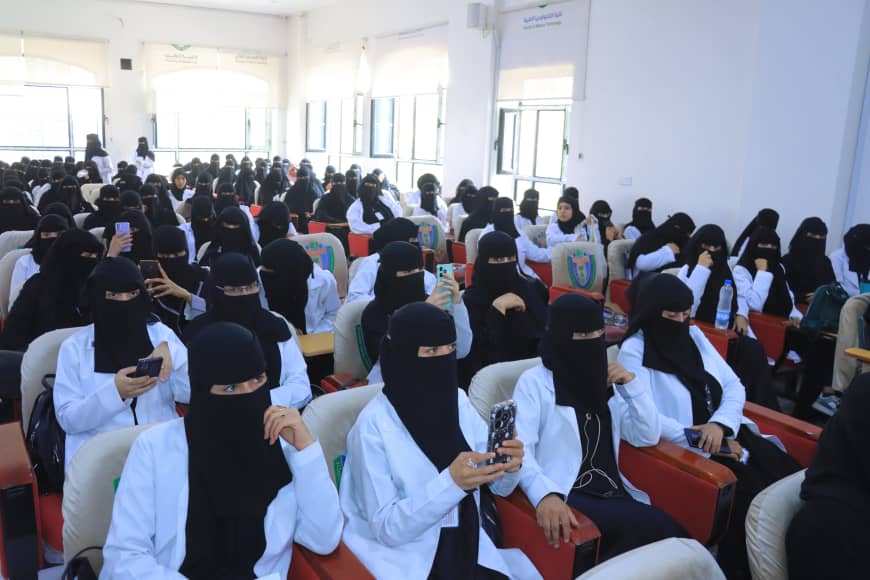 Group of women in white coats and black niqabs seated in a bright lecture hall, many looking at their phones.