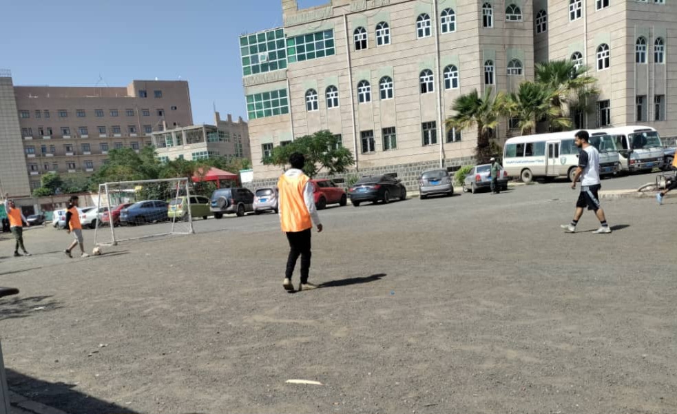 Person in an orange vest stands on a dusty field as others play soccer near a small goal, with a large campus-style building and parked cars in the background.