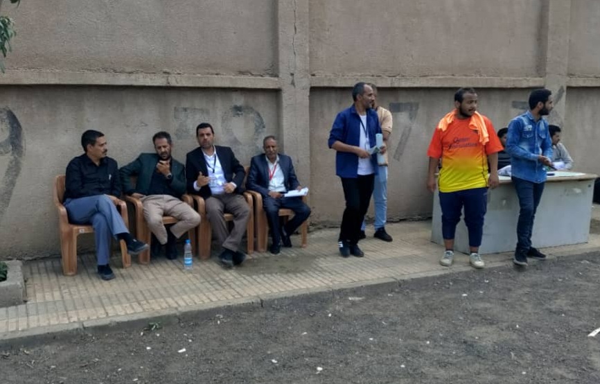 Group of men seated on plastic chairs outdoors beside a concrete wall, some with lanyards, as others stand nearby.