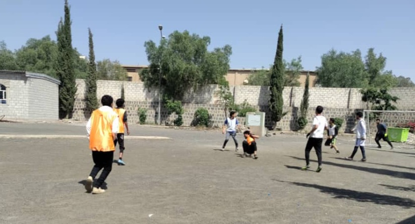 Children playing a game on a dusty outdoor field; some wear orange vests, with a stone wall and trees in the background.