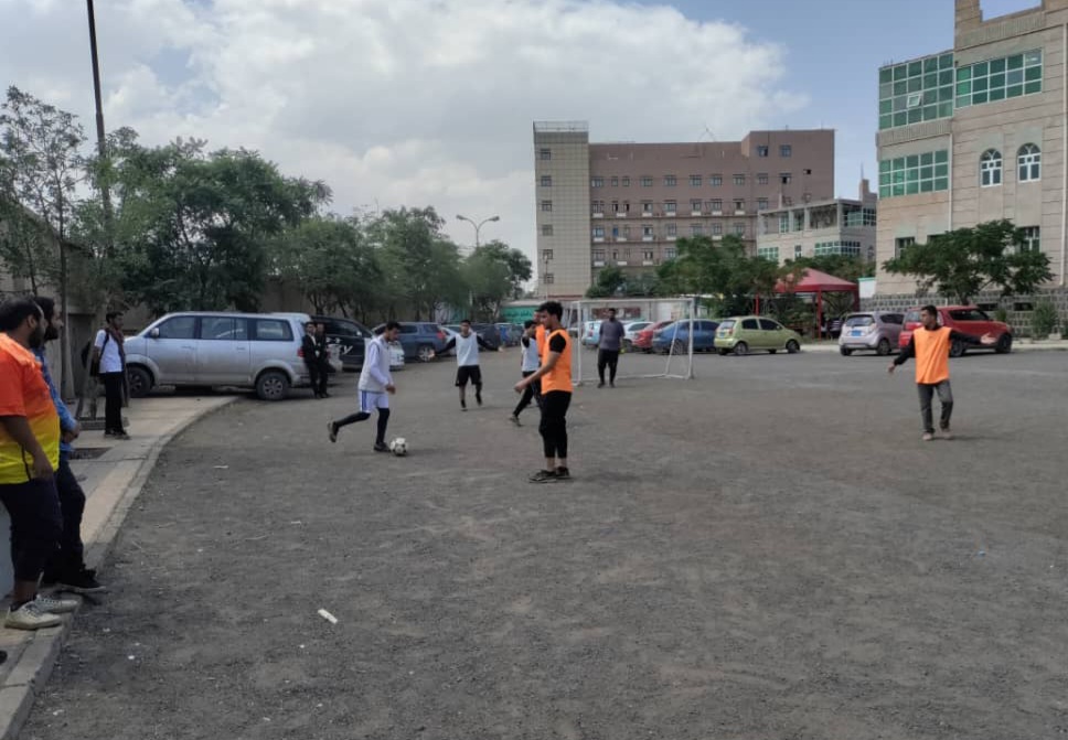 People playing soccer on a gravel field beside parked cars, with players wearing orange vests and white shirts against an urban backdrop.