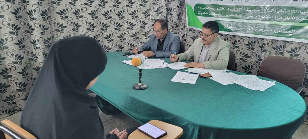 Woman in a hijab faces two men at a green-covered table, signing documents during a press conference; a microphone sits between them, with a banner in the background.