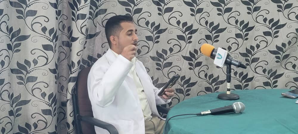 Man in a white lab coat sits at a table with a microphone and a phone, as if in an interview or press setup.