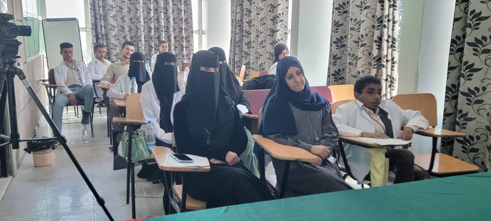 Classroom scene with students seated at desks, including women in traditional dress, listening attentively as a camera records from the side.