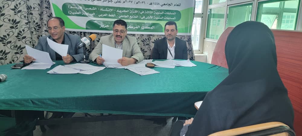 Three men seated behind a green-covered table reading documents during a press conference, with a woman in a black hijab facing them and microphones on the table in front of them.