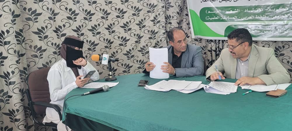 Panel discussion at a press conference: three men sit at a green table with papers; left wears white robe and black niqab, middle in gray suit, right in beige suit; banner with Arabic text behind.