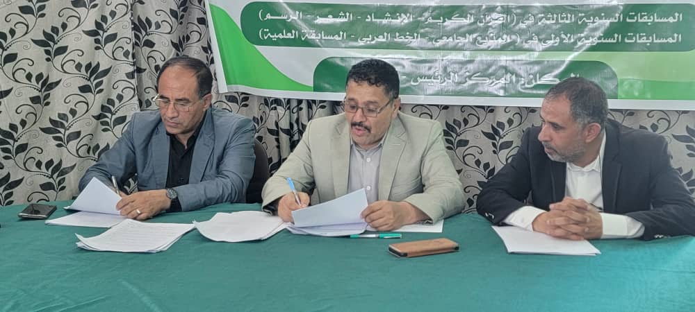 Three men sit at a green table signing and reviewing documents as a banner with Arabic text hangs behind them.