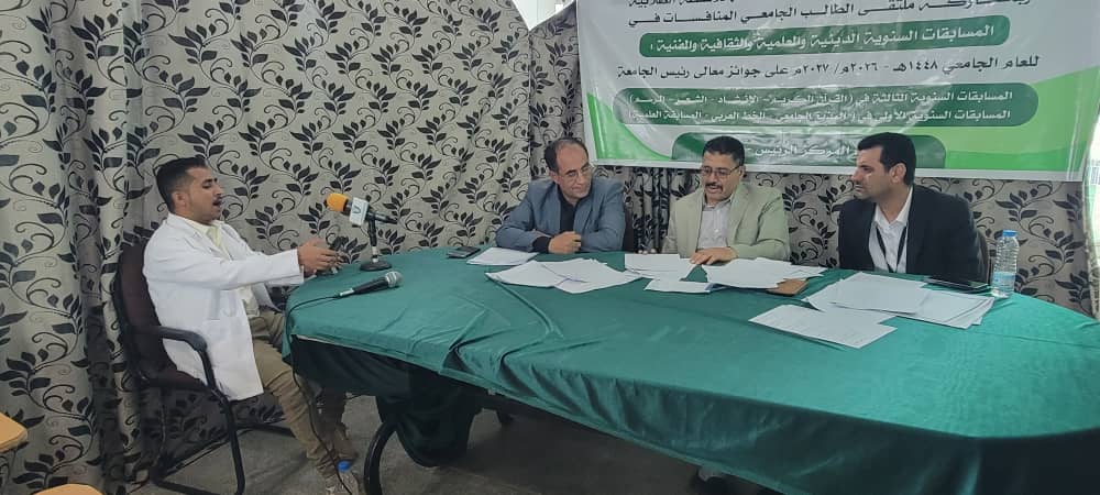 Four men sit at a green-clothed table during a panel discussion; one man in a white coat speaks into a microphone, with papers on the table and a banner with Arabic text in the background.