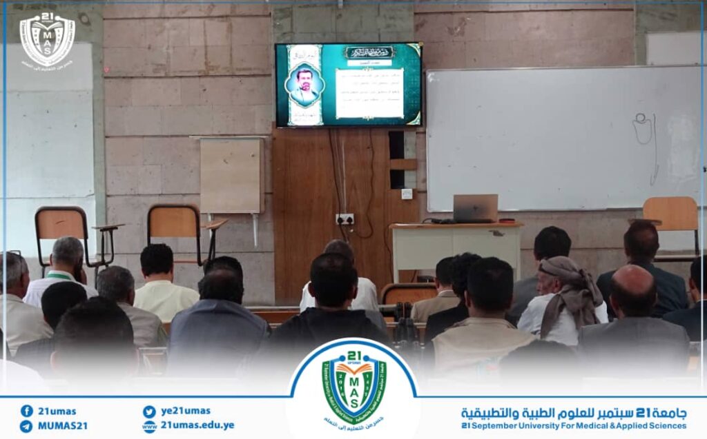 Lecture hall with adults seated facing a presentation on a mounted screen; a desk with a laptop and whiteboard backdrop behind. A university banner and logo appear along the bottom edge.