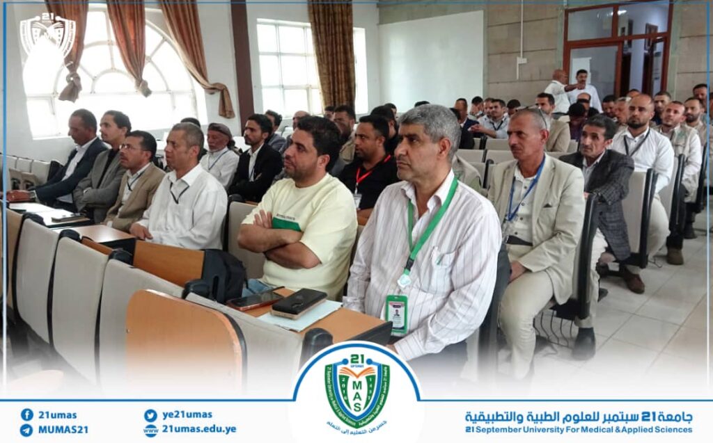 Audience of men in a lecture hall listening to a presentation; front-row man wears a green lanyard.