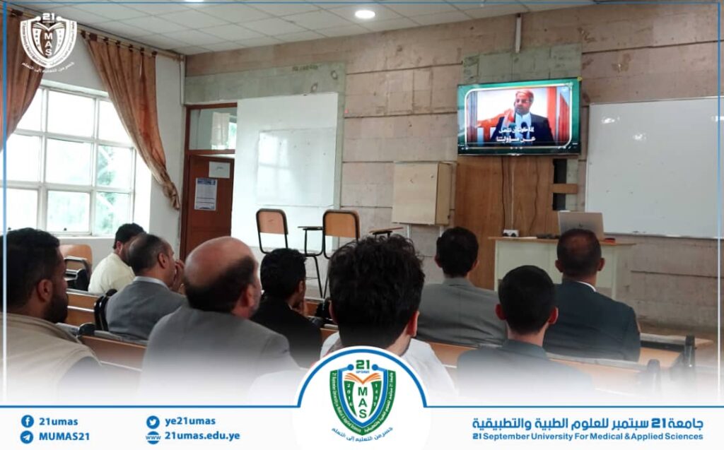 Classroom audience watching a lecture on a TV screen in a university hall, with 21mas logo and Arabic text on screen.