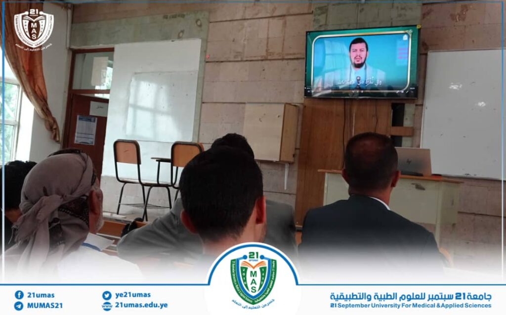 Students in a classroom watching a video lecture on a wall-mounted TV, with university branding visible at the bottom edge