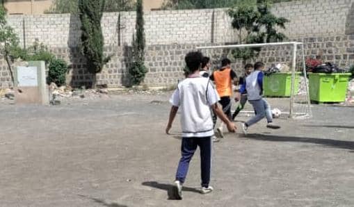 Children playing soccer on an outdoor dirt field near a goal, with a brick wall and green dumpsters in the background.