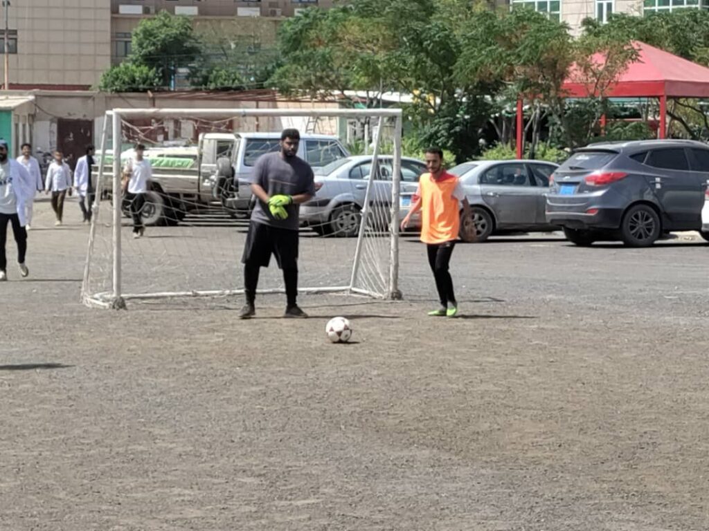Two men stand near a small soccer goal on a dusty field, with a ball between them; one is a goalkeeper wearing neon gloves and a gray shirt, the other in an orange shirt.