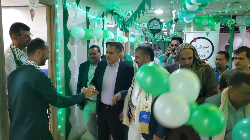 Group of men in a hallway birthday/celebration setup, shaking hands under green balloon decorations and streamers.