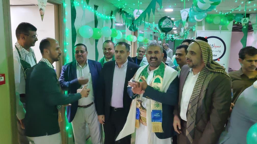 Group of men in a festively decorated hallway with green lights and balloons, chatting and smiling at a celebration.