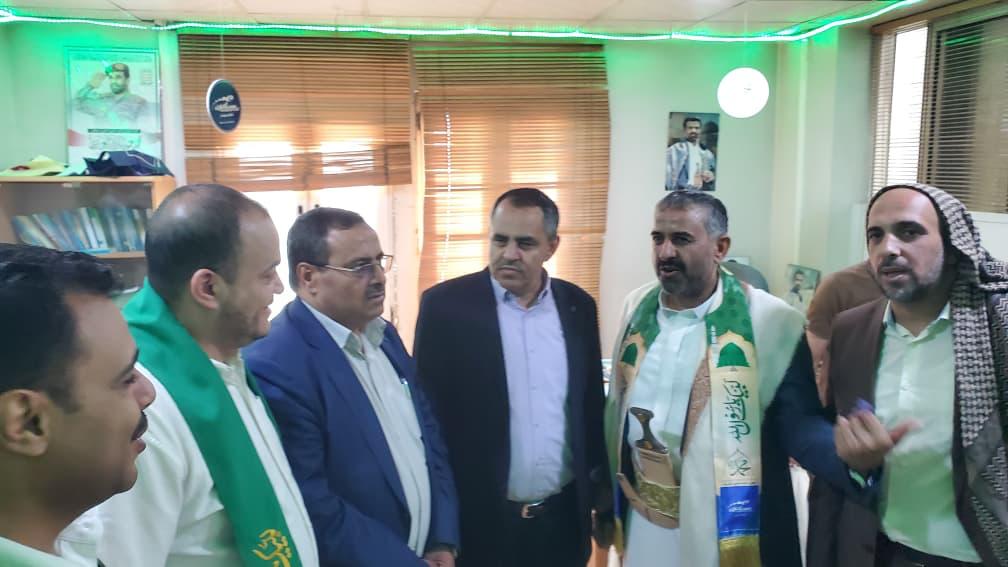 Group of six men standing in a bright room, some wearing green ceremonial scarves, engaged in conversation during a formal gathering.