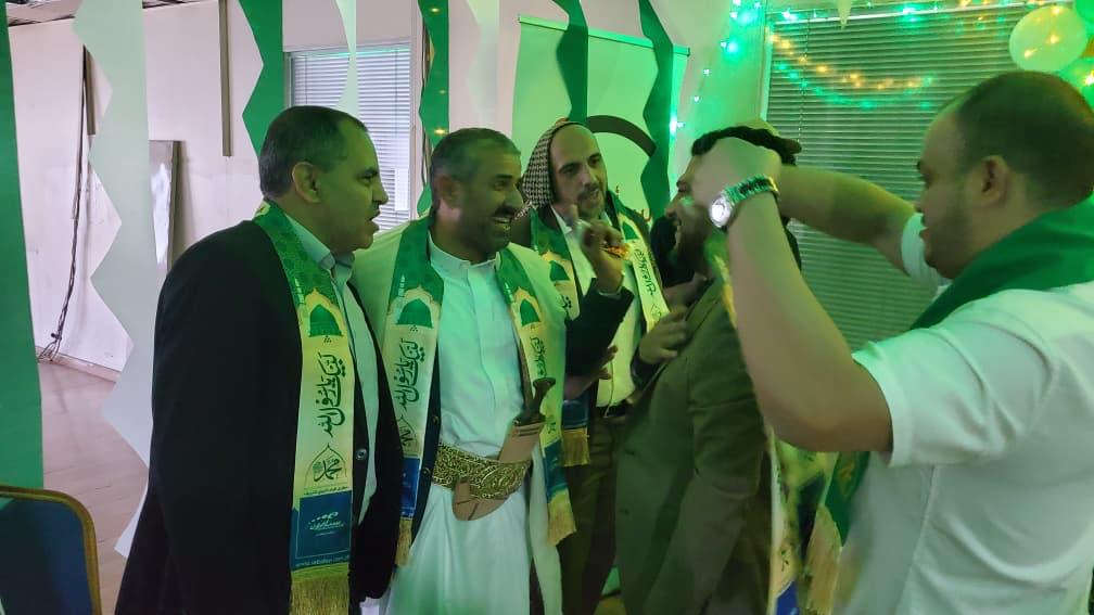 Group of men at a festive event wearing green sashes, smiling as one man pins a medal onto another.