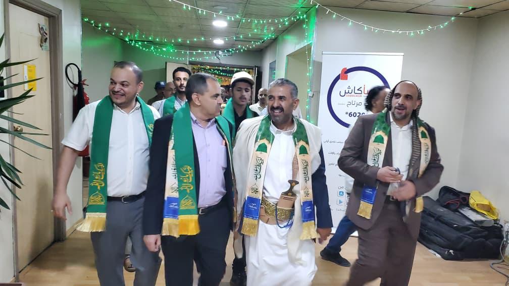 Group of men wearing green and gold ceremonial sashes walk through a hallway decorated with green string lights.