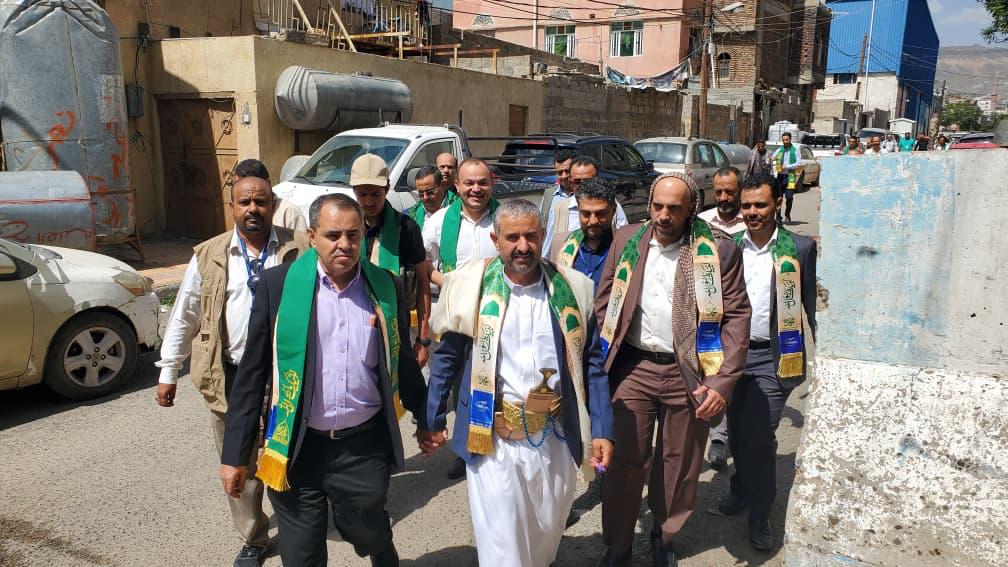 A group of men wearing green-and-gold scarves walk together down a sunlit urban street, appearing to form a delegation or procession.