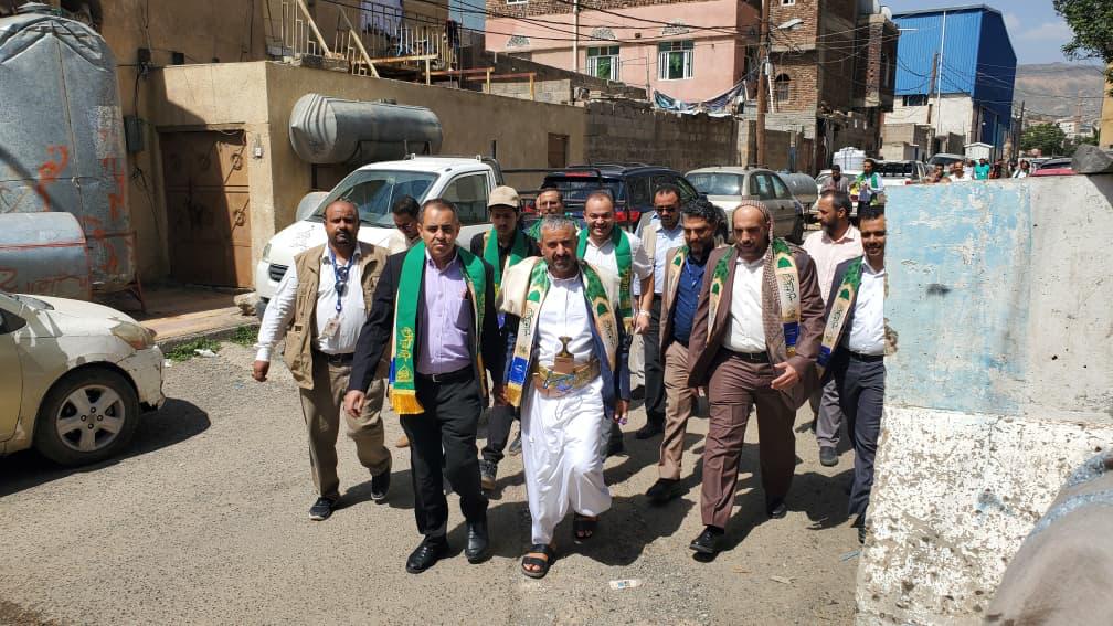 Group of men wearing green ceremonial scarves walking together down a sunlit urban street with parked cars and buildings in the background, forming a procession.