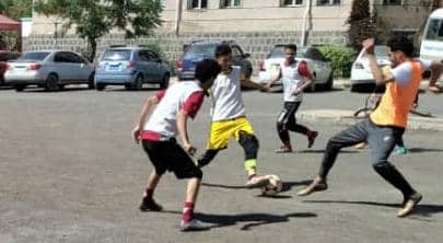 Group of five people playing casual soccer in a paved parking lot, vying for the ball across the driveway area.