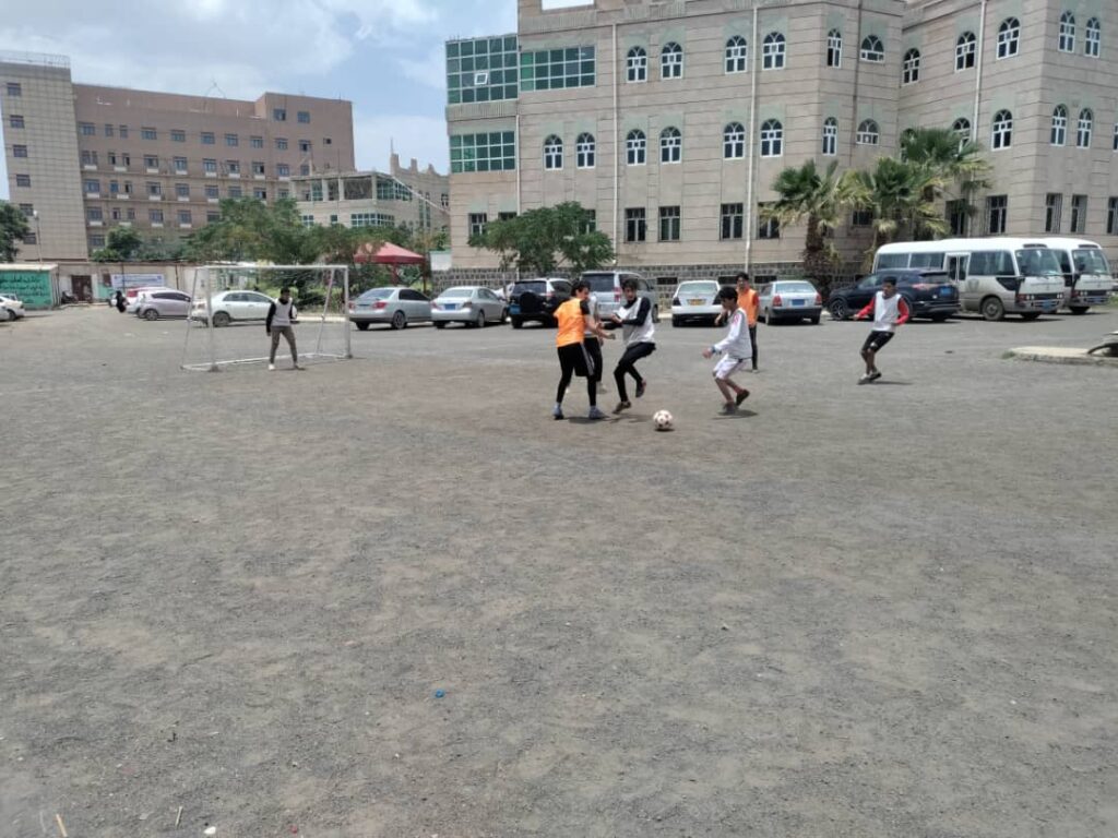 Group of students playing soccer on a dusty field near a portable goal, with cars and a large campus building in the background.