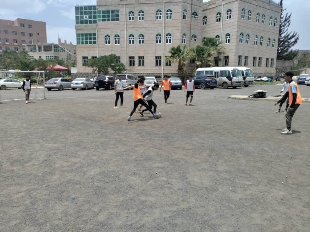 Group of young people playing soccer on a gravel lot in front of a large stone building, with some players in orange pinnies and cars parked nearby.