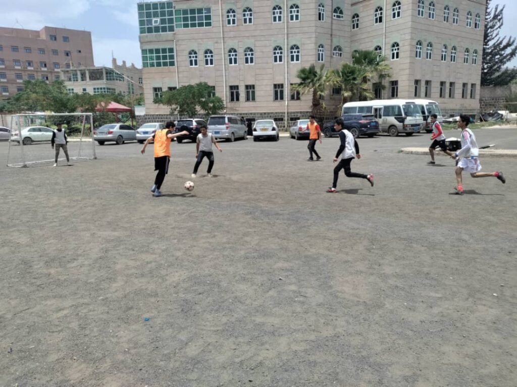 Group of youths playing soccer on a gravel field in front of a large campus-style building; a small goal is on the left and players chase the ball nearby.