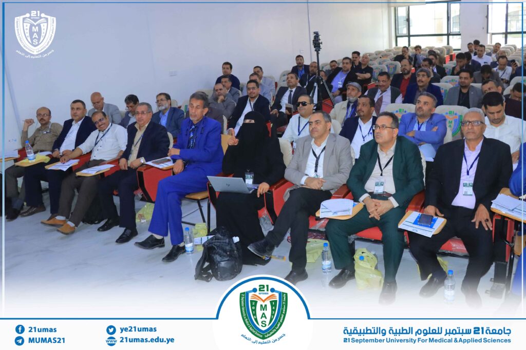 Audience at a university conference, rows of professionals listening in a bright auditorium with a large logo banner in the foreground.