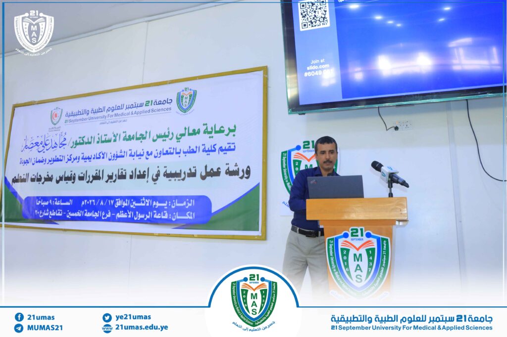 Man stands at a wooden podium giving a talk at a university event, with a large banner in Arabic and the 21 September University for Medical & Applied Sciences logo visible in the background.