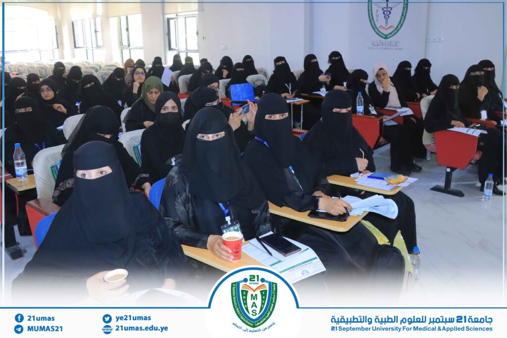 Large lecture hall filled with women in black abayas and hijabs seated at desks, taking notes during a seminar; university logo visible on the wall.