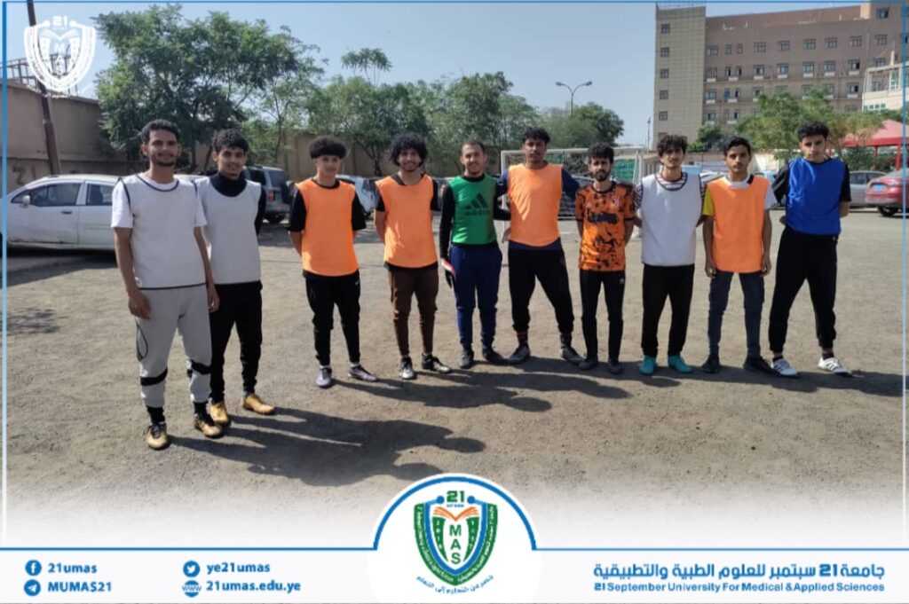 Eleven young men stand in a line on a dirt field, wearing training bibs, posing for a group photo with a university crest visible along the bottom edge.
