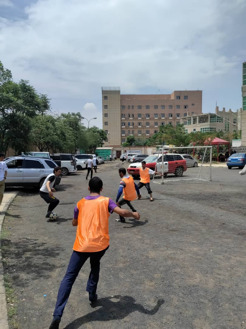Group of young men playing soccer in a parking lot, wearing orange vests, with a goal in the background and urban buildings nearby.