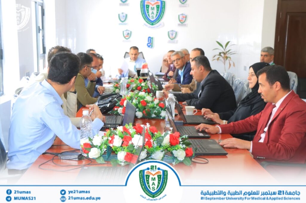 Group of professionals around a conference table with laptops and a flower centerpiece during a formal meeting in a university setting.