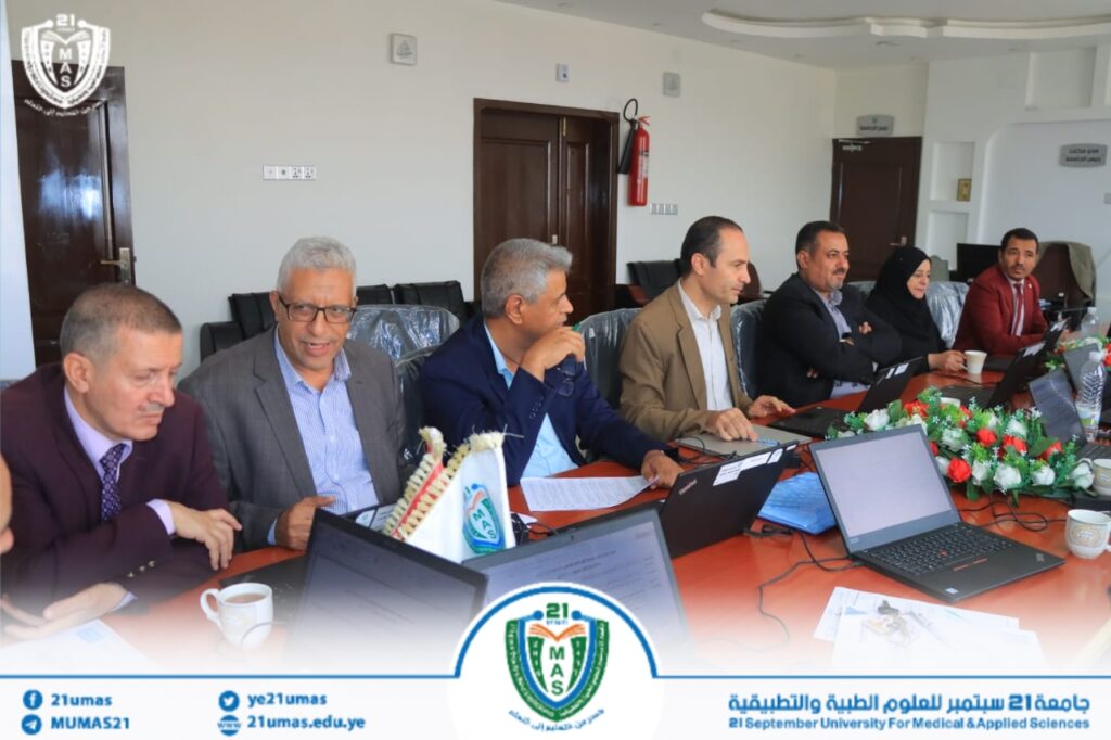 Group of professionals in a formal meeting around a conference table with laptops; university banner and crest visible at bottom.