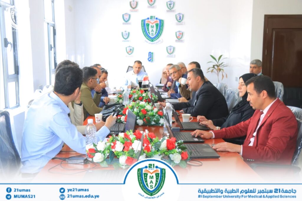 Group of professionals seated around a long conference table with laptops, in a formal meeting at a university room with emblem banners on the wall in the background.