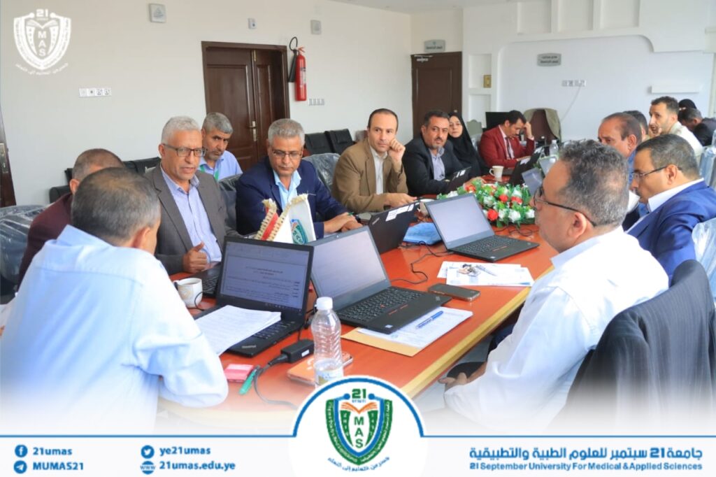 Group of professionals around a conference table with laptops during a formal university meeting, banner and logo in the background.