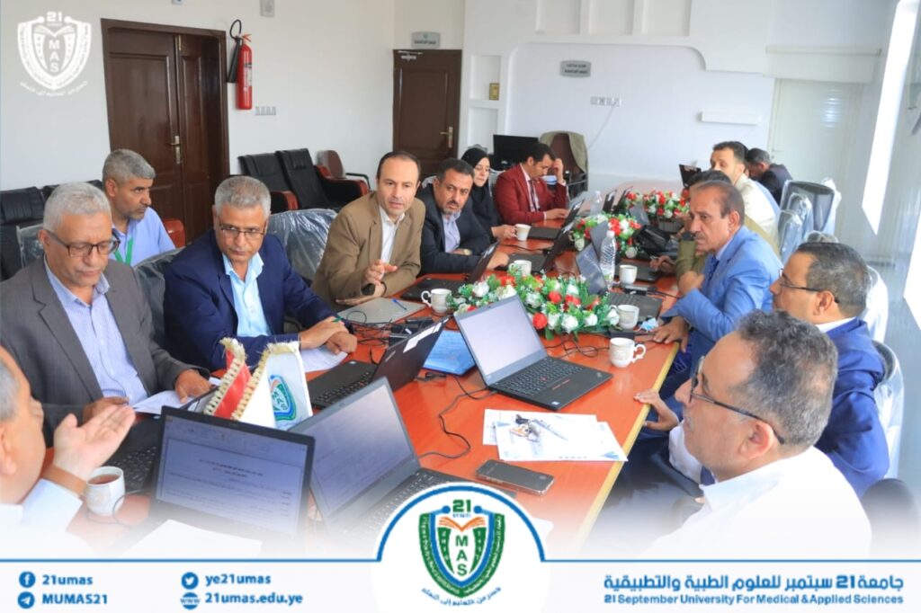 Professionals in a conference room gathered around a long table with laptops, papers, and cups for a meeting, university banner visible at the bottom edge.